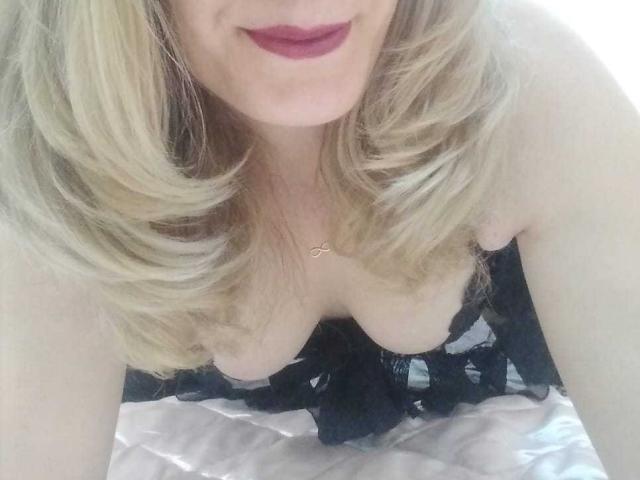 Una donna bionda, con i capelli mossi, rossetto rosso e un corpetto di pizzo nero, che mostra il seno, è china in avanti e indossa una collana d'oro..