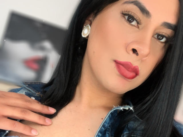 Sara Winson tem a pele morena, cabelo preto, batom vermelho e está vestindo uma jaqueta jeans com a mão no peito. Ela usa um brinco de pedra preciosa e olha para a câmera..
