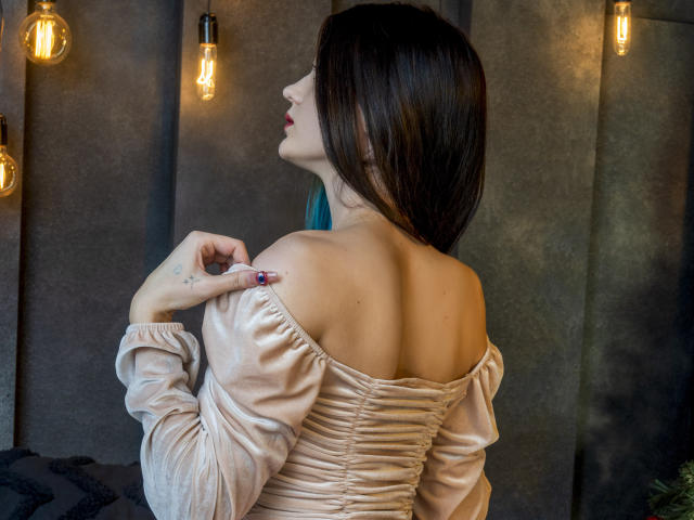 Nicole, cheveux bruns foncés, tournée vers la gauche, portant un haut beige à une épaule, laissant apparaître le haut du dos, tenant une bretelle, avec fesses et tatouage sur le dos visibles, sous des ampoules lumineuses chaudes..