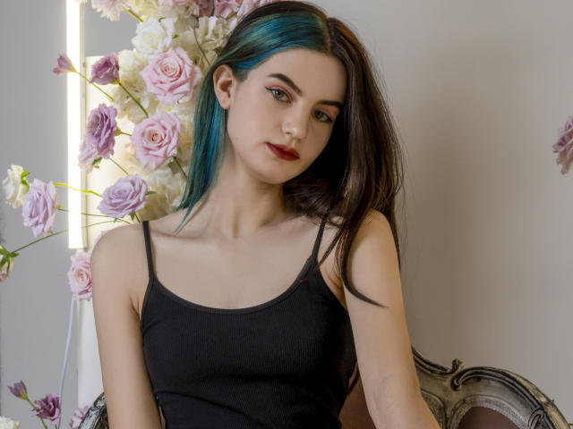 NicoleKissYou met turquoise blauwe highlights in donkerbruin haar, gekleed in een zwarte tanktop, zittend met roze en witte rozen op de achtergrond, kijkend naar de camera..