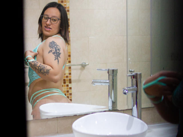 NatalieHarris with glasses, green lace bra, and matching strappy panties, showing a rose tattoo on her right arm, standing in a bathroom, looking at herself in the mirror.