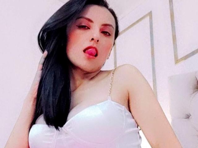 NataliaCamp, com cabelo preto, maquiagem rosa e batom vermelho, língua de fora, vestindo uma blusa branca, sem alças, estilo corset, mostrando seus seios. Ela está tocando o cabelo..