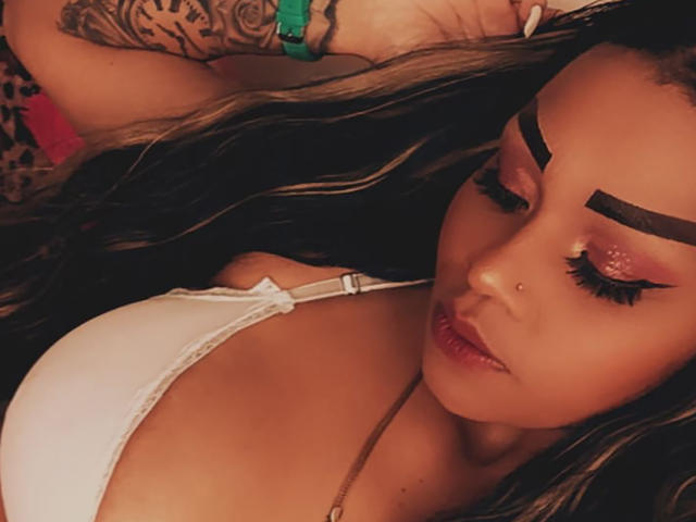 LinaaPrincess with long, wavy hair, wearing a white bra, has thick, dark eyebrows, glittery pink eyeshadow, and a nose ring, lying down with a tattooed arm.