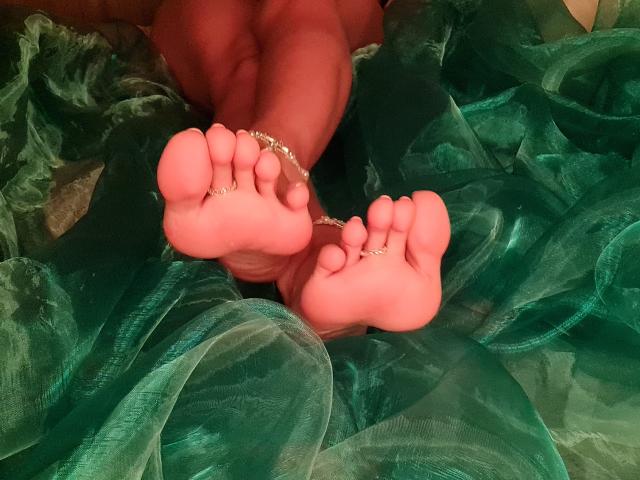 De roze zolen van AnaMariaDiaz, met zilveren ringetjes aan de tenen, haar voeten gekruist, liggen op een groen, gekreukeld doek. Haar benen zijn licht zichtbaar op de achtergrond..