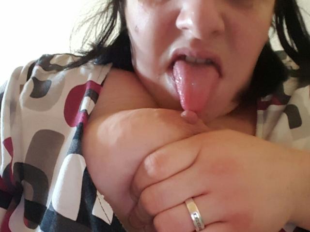 MimiSammy, mit herausgesteckter Zunge, leckt ihre großen, nackten Brüste. Sie hält sie mit ihrer Hand, trägt einen Ring und hat ein gemustertes Hemd an..