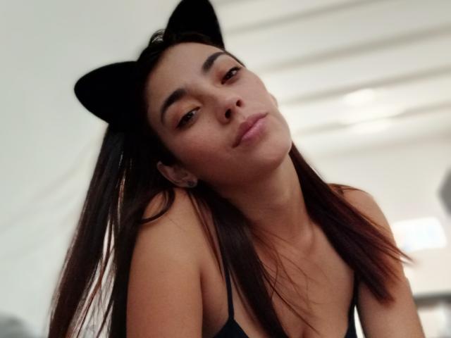 HannaAndHarri con orecchie a forma di gatto nero, capelli castani, un leggero broncio, indossa una maglietta nera, guarda in basso, naso piccolo, pelle chiara, piercing alle orecchie, luce soffusa, casual, sexy, sicura di sé..