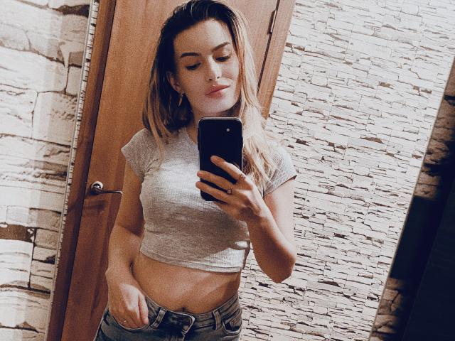 Carol Rose, avec ses cheveux blonds, portant un haut court gris qui laisse entrevoir son ventre, prend un selfie devant un miroir, tenant un téléphone noir, et se tient devant une porte en bois..