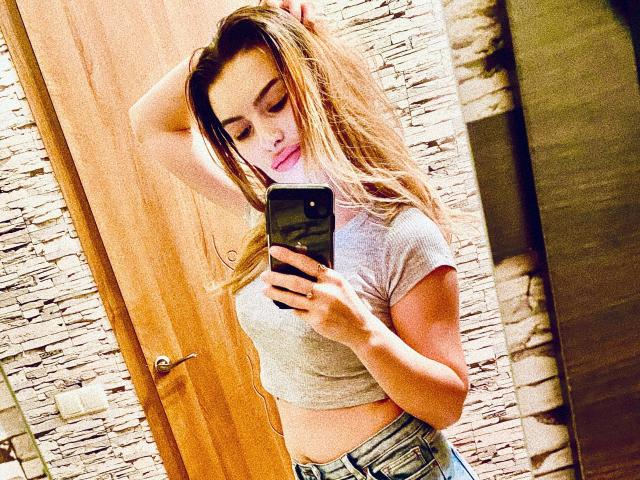 Carol Rose mit langen, braunen Haaren, die ein graues Crop-Top und blaue Jeans trägt, macht ein Selfie im Spiegel, wobei sie eine Hand in ihrem Haar hat und ihre kleinen Brüste und ihr flaches Gesäß zeigt..