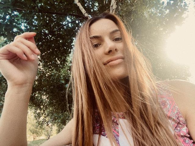 Carol Rose, con cabello castaño largo, mirando a la cámara, sosteniendo una hoja, vistiendo una blusa rosa con estampado, luz solar en el fondo, una ligera sonrisa, luz natural, ambiente casual al aire libre..