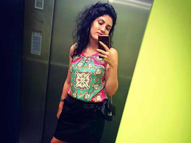 AddictedToUTS with curly black hair, wearing a colorful, patterned top and black skirt, holding a phone, standing in an elevator, showing off her tits and ass.