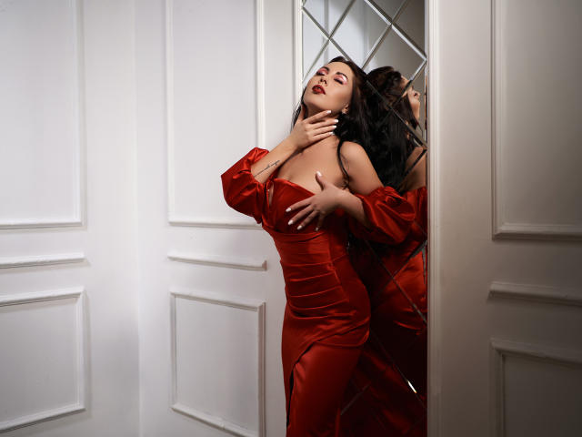 NinaTender in a shiny red dress, touching her tits, with her reflection in a mirror, hand on her neck, black wavy hair, white wall, and door in the background.