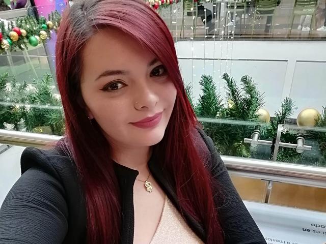 KatherinLatin with red hair, black jacket, and pink lipstick, smiling at the camera, showing off her nose ring and cute face, wearing a gold necklace.