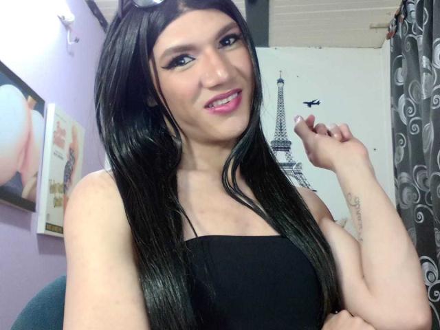 LunaGransTs with long black hair, wearing a black strapless top, smiling with pink lipstick, holding a finger to her cheek, showing a tattoo on her right arm.