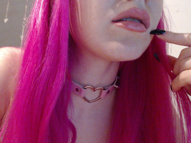 SweetyPieX con i capelli rosa, lo smalto nero alle unghie e una collana con un cuore rosa, che si morde un dito, mostrando le sue labbra rosa leggermente dischiuse e la pelle pallida..