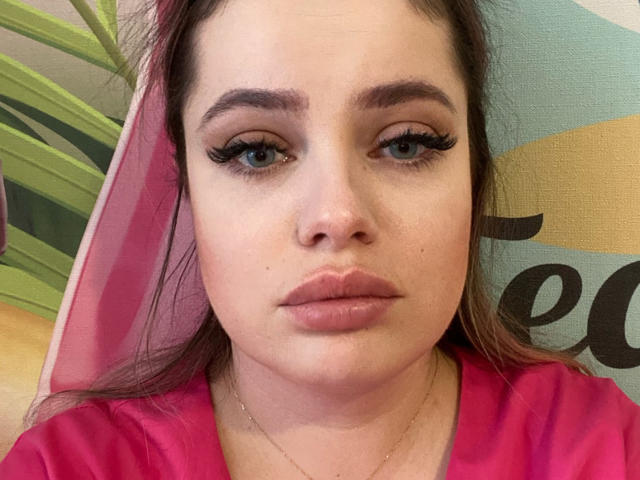 Marizel with pink lipstick, full eyebrows, long eyelashes, and a pink shirt, has a neutral expression, light skin, and brown hair. She's wearing a thin gold necklace.