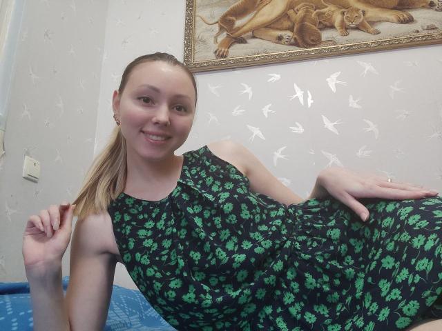 AliceSmiles, with blonde ponytail, smiling, wearing a green floral dress, reclining on a blue bed, right hand on hip, left hand near head, showing small tits and slim ass.