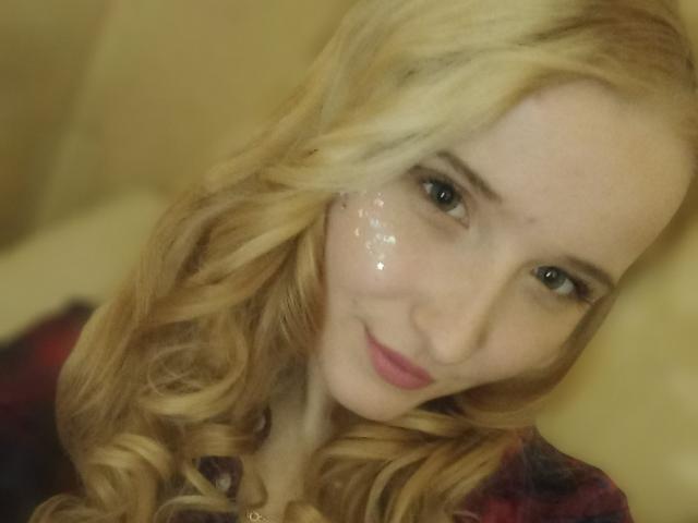 MelannieX with blonde curls, shiny face, and pink lips, smiling softly, wearing a red and black plaid shirt, in a blurred photograph.