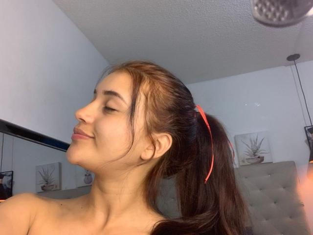 HelenTay with brown ponytail, red hair tie, smiling, eyes closed, nude, showing her tits, fair skin, white wall, grey tufted chair, ceiling light, subtle makeup.