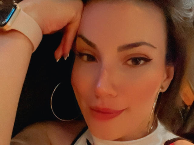 Liliysweet hat ein süßes Gesicht mit dunklen Augenbrauen, einem Eyeliner im Winged-Stil und pinkem Lippenstift. Sie trägt ein pinkes Armband und große Ohrringe im Hula-Hoop-Stil und hat ein leichtes Lächeln..