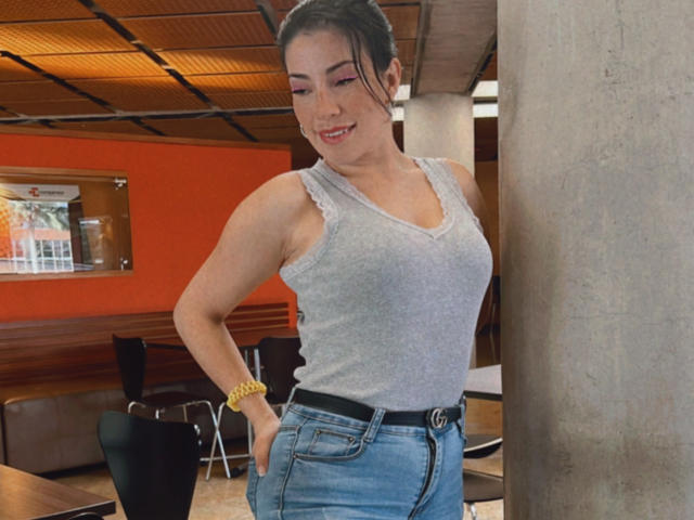 Liliysweet met een schattige glimlach, een grijze tanktop die kleine borsten laat zien, blauwe jeans, een gele armband, handen in de zakken, staand naast een betonnen pilaar in een moderne kamer..