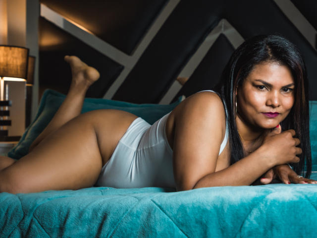 Une femme, AmberColt, avec une peau brune, des cheveux noirs et du rouge à lèvres violet, allongée sur un lit bleu turquoise, portant une combinaison blanche, montrant ses fesses et regardant l'appareil photo..