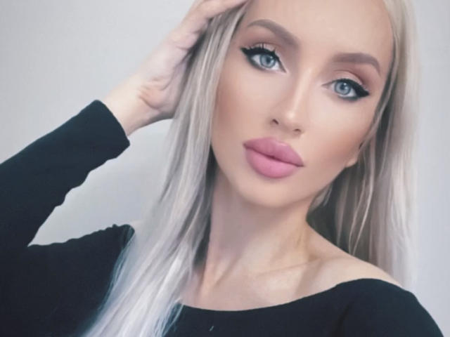 GoddessZora a de longs cheveux blonds platine, des yeux bleus, du rouge à lèvres rose, et elle porte un haut noir à une épaule avec une main dans ses cheveux. Ses seins ne sont pas visibles..