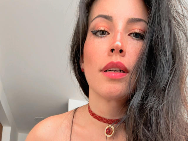 Il viso di Liliysweet, con rossetto rosso, eyeliner nero e una collana con un ciondolo a forma di cuore. Ha lunghi capelli neri e un leggero sorriso..