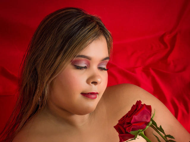 EvaMadison with red eyes, pink eyeshadow, and red lipstick, holding a red rose, has brown hair, and is topless, against a red background. Her tits are slightly visible.