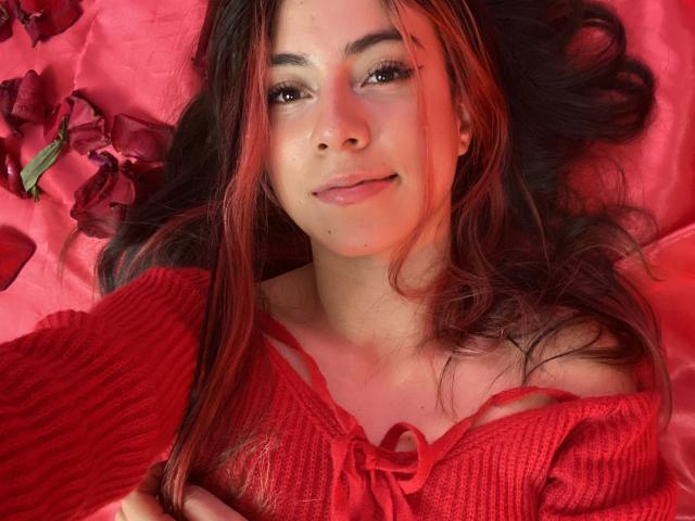 JuliaGarciax with red ribbed shirt, small tits, lying on pink bed, dark wavy hair, subtle smile, red petals, soft lighting, brown eyes, natural makeup, slightly flushed face.