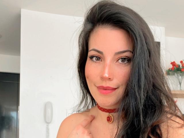 Liliysweet tiene el pelo largo y oscuro, lápiz labial rojo y un collar con un colgante rojo que tiene una gema roja. Está sonriendo y tiene una mano cerca del cuello. No se ven los senos..
