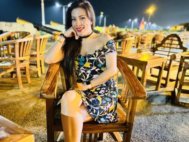 Liliysweet with long dark hair, smiling, wearing an off-shoulder floral dress, black choker, and tan stockings, sitting on a wooden chair, hand on cheek, at night.
