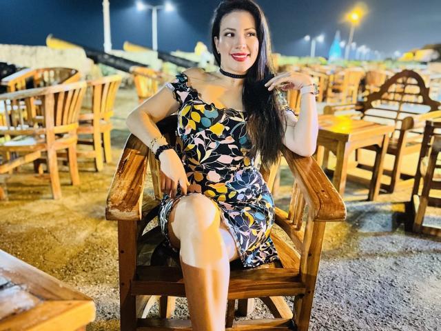 Liliysweet with long black hair, red lipstick, and floral dress, sitting on wooden chair, leg crossed, smiling, black choker, tattoos on wrist, night setting, streetlights in background.
