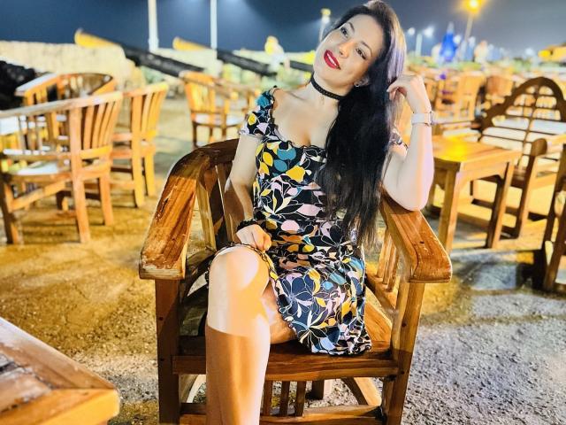 Liliysweet, sitting on a wooden chair, wearing a floral dress, showing her tits, with a black choker, long black hair, red lipstick, and a confident smile.