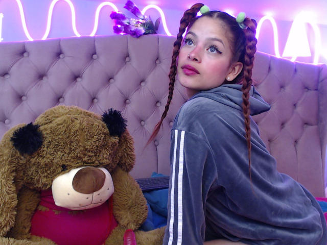 ShantalSummer with braided pigtails, green hair ties, gray hoodie, pink lipstick, and blue eyes, sitting next to a brown teddy bear with a red shirt.
