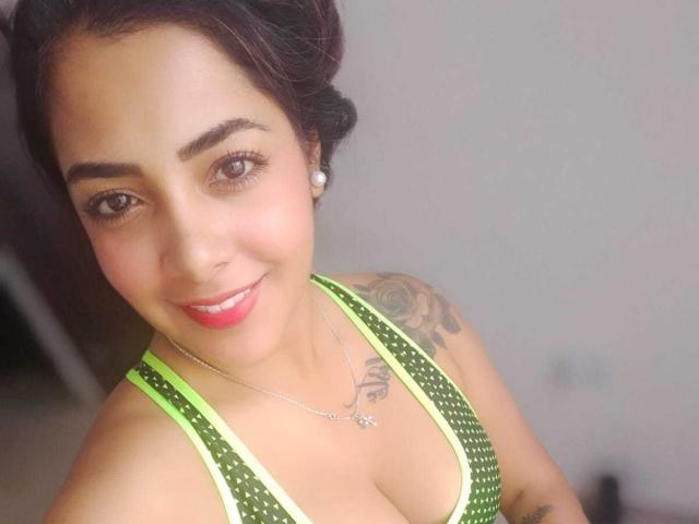 Briana Escobar con rossetto rosso, top a pois verdi che mostra il seno, tatuaggio sulla spalla destra, orecchini, che sorride, occhi marroni, capelli neri raccolti in uno chignon e una collana..