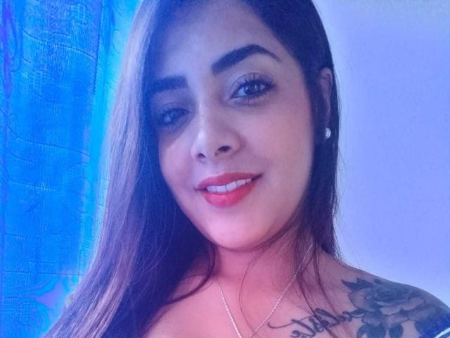 Briana Escobar, com cabelos castanhos longos, sobrancelhas grossas, batom vermelho, sorrindo, mostrando uma tatuagem no ombro direito, usando um colar de prata e brincos pequenos..