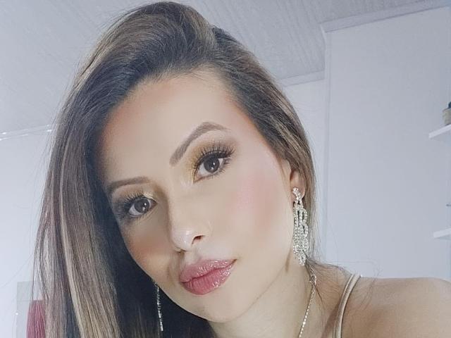 HannaRobertss has brown hair, shiny pink lips, and wears glittery eyeliner with long lashes. She's sporting silver earrings and a necklace, with a slightly pouty expression.