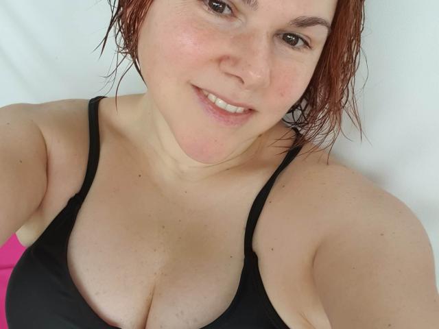 SportyLove with red wet hair, smiling, wearing a black bra that shows her big boobs, fair skin, and brown eyes. Her pink underwear is slightly visible.