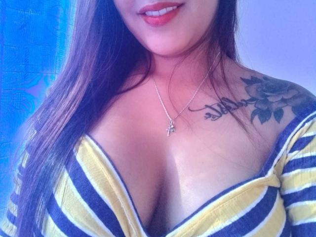 Briana Escobar, com seios grandes, vestindo uma camisa listrada amarela e preta, colar com cruz, tatuagem no ombro direito, cabelo castanho comprido, sorrindo, batom vermelho..