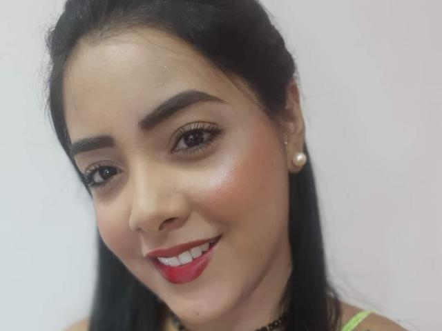 Briana Escobar tiene ojos marrón oscuro, cabello negro, cejas pobladas, lápiz labial rojo y lleva aretes de perlas. Está sonriendo, tiene la piel brillante y un collar negro..