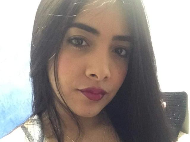 Briana Escobar tiene el cabello largo y negro, cejas oscuras, ojos marrones y lápiz labial rojo. Su rostro está centrado en la imagen, mostrando una ligera sonrisa con una expresión neutra..