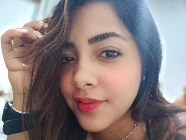 O rosto de Briana Escobar, cabelo castanho escuro ondulado, sobrancelhas grossas, batom vermelho, sorriso discreto, segurando o cabelo, piercing no nariz, colar de prata, luz suave, fofa, atraente..