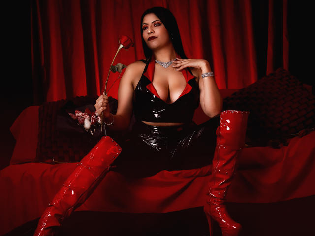 Kamile Nilsen, con botas rojas, top de látex negro y escote, sosteniendo una rosa, sentada en un sofá rojo, cortinas rojo oscuro, almohadas negras y rojas, iluminación rojo oscuro..