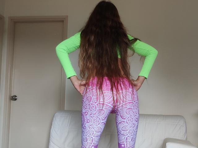 Honytsoi mit langen, braunen Haaren, einem grünen Oberteil und violett/rosa gemusterten Hosen, die Hände an den Hüften, steht vor einer weißen Tür und einem Sofa..