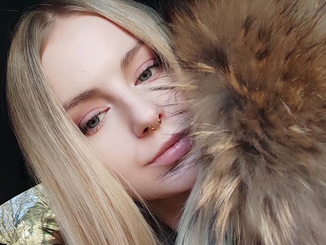 EvaYuga, com cabelo loiro, olhos verdes e um piercing no nariz, está tirando uma selfie com um grande animal peludo marrom e preto. O animal está encostado na bochecha direita dela..