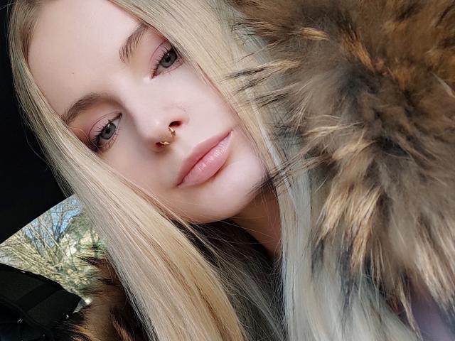 Le visage d'EvaYuga, avec ses cheveux blonds, son anneau de nez et son maquillage discret, portant une veste bordée de fourrure, légèrement inclinée, avec la fourrure au premier plan..