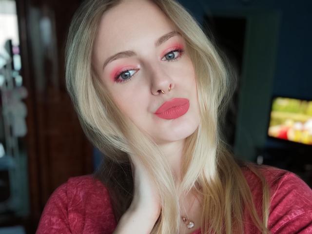 EvaYuga, con cabello rubio, maquillaje de ojos rosa, lápiz labial rojo, piercing en la nariz y una camisa rosa, tocándose el cuello, ojos azules, con una ligera sonrisa, y llevando un collar..