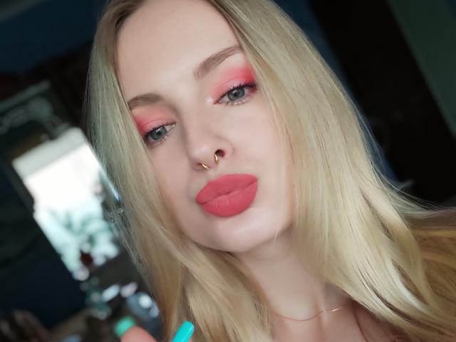 EvaYuga met blond haar, roze oogschaduw, rode lipstick, een neusring en blauwe nagels, kijkt in de camera met lichtjes open mond, zacht licht op het gezicht, een zachte uitdrukking..