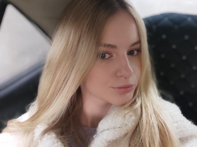 EvaYuga mit blonden Haaren, einem Nasenring und einer weißen, flauschigen Jacke, die leicht zur Seite schaut und ein leichtes Lächeln trägt. Ihre Augen sind braun und ihre Lippen sind rosa..