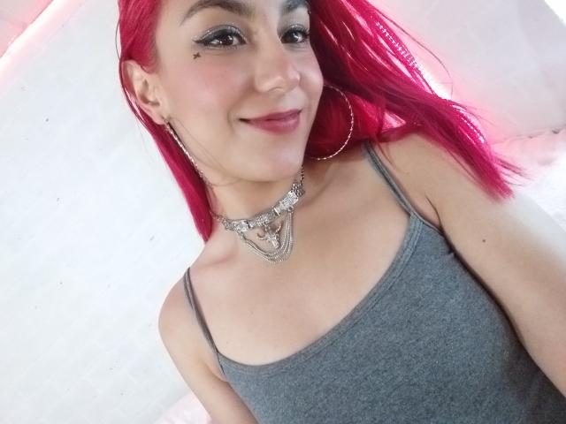 AmyStreetwise69 with bright pink hair, wearing a grey tank top, hoop earrings, and a fancy necklace, smiling at the camera, showing her cute face and small tits.